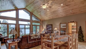 A spacious living room with a large wooden dining table, a sofa, and a fireplace. There are large windows offering a view of the outdoors.