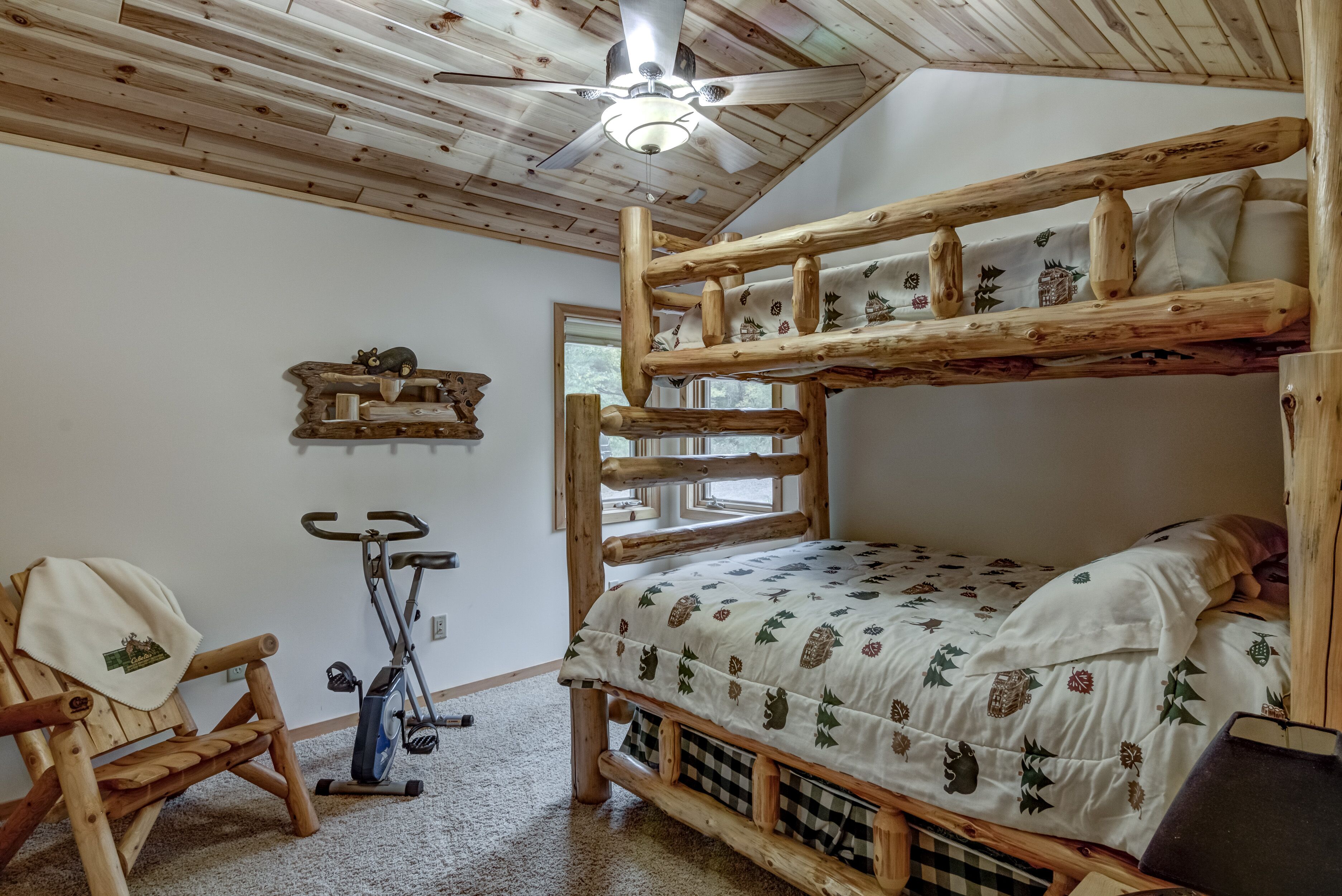 Un letto a castello con copripiumino floreale, una sedia in legno, una cyclette e una ventola a soffitto in una stanza con pareti in legno e una finestra.