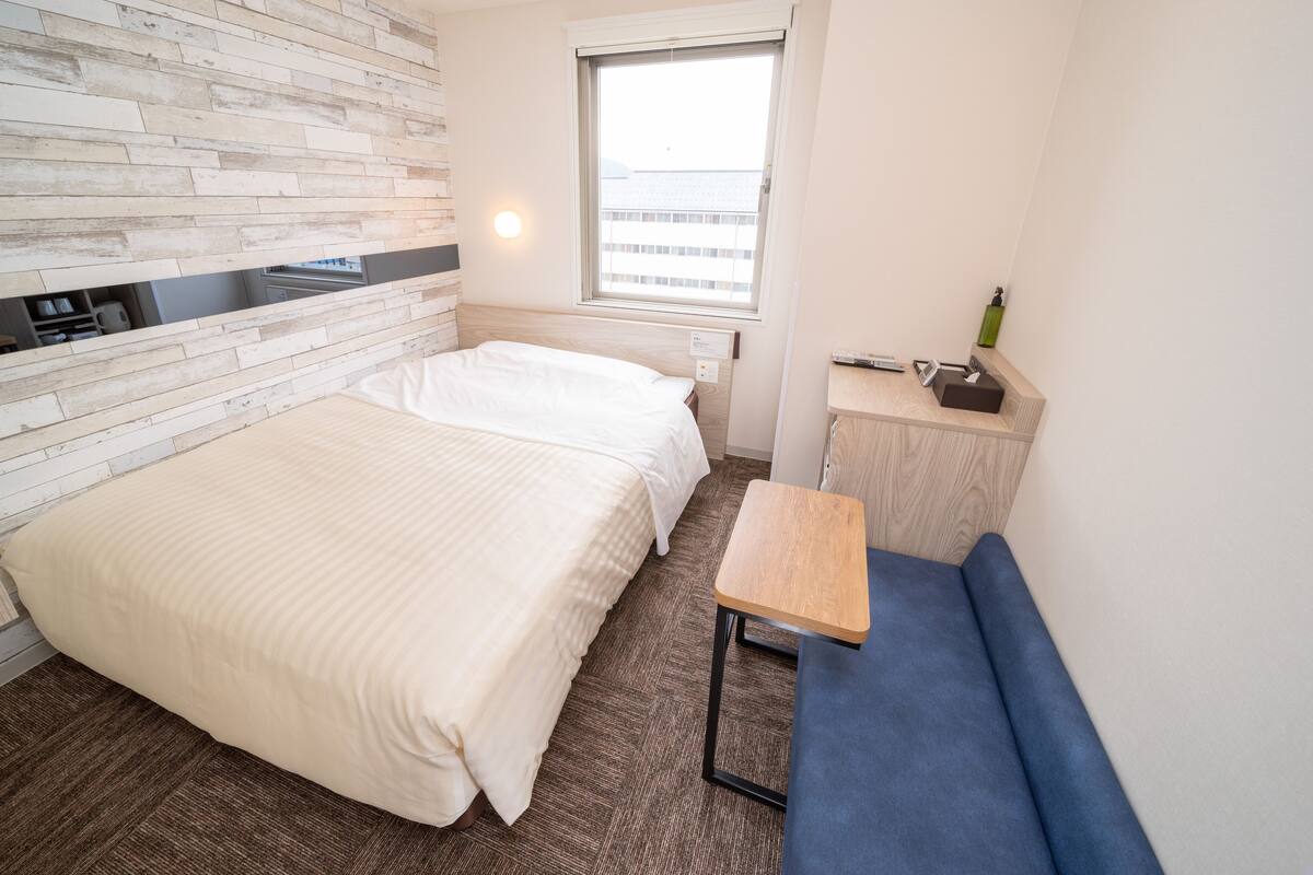 A hotel room with a bed, a small desk, a bench, a window, and a wall with a wooden pattern.