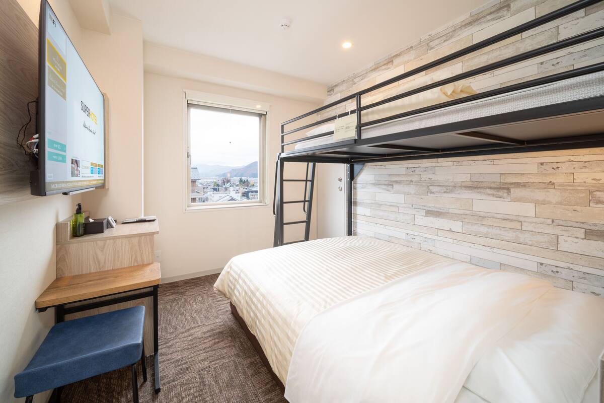 A compact hotel room with a bunk bed, a desk with a chair, a flat-screen TV, and a window offering a city view.