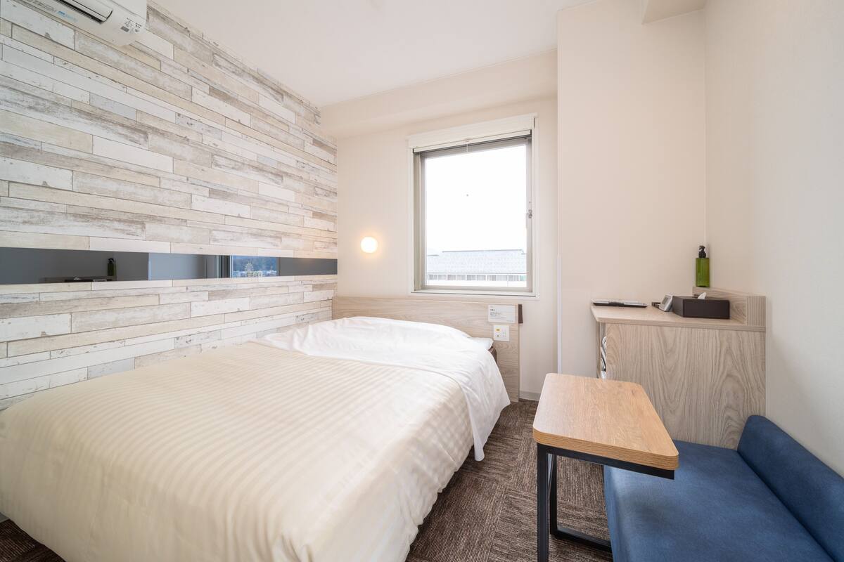 A hotel room with two beds, a small table, a blue sofa, a window with a view, and a wall-mounted air conditioner.