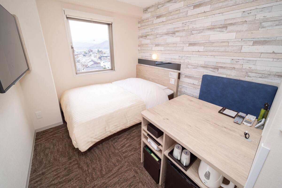 A compact hotel room with a single bed, a desk with a chair, a small shelf with toiletries, a window with a city view, and a wall with a textured pattern.