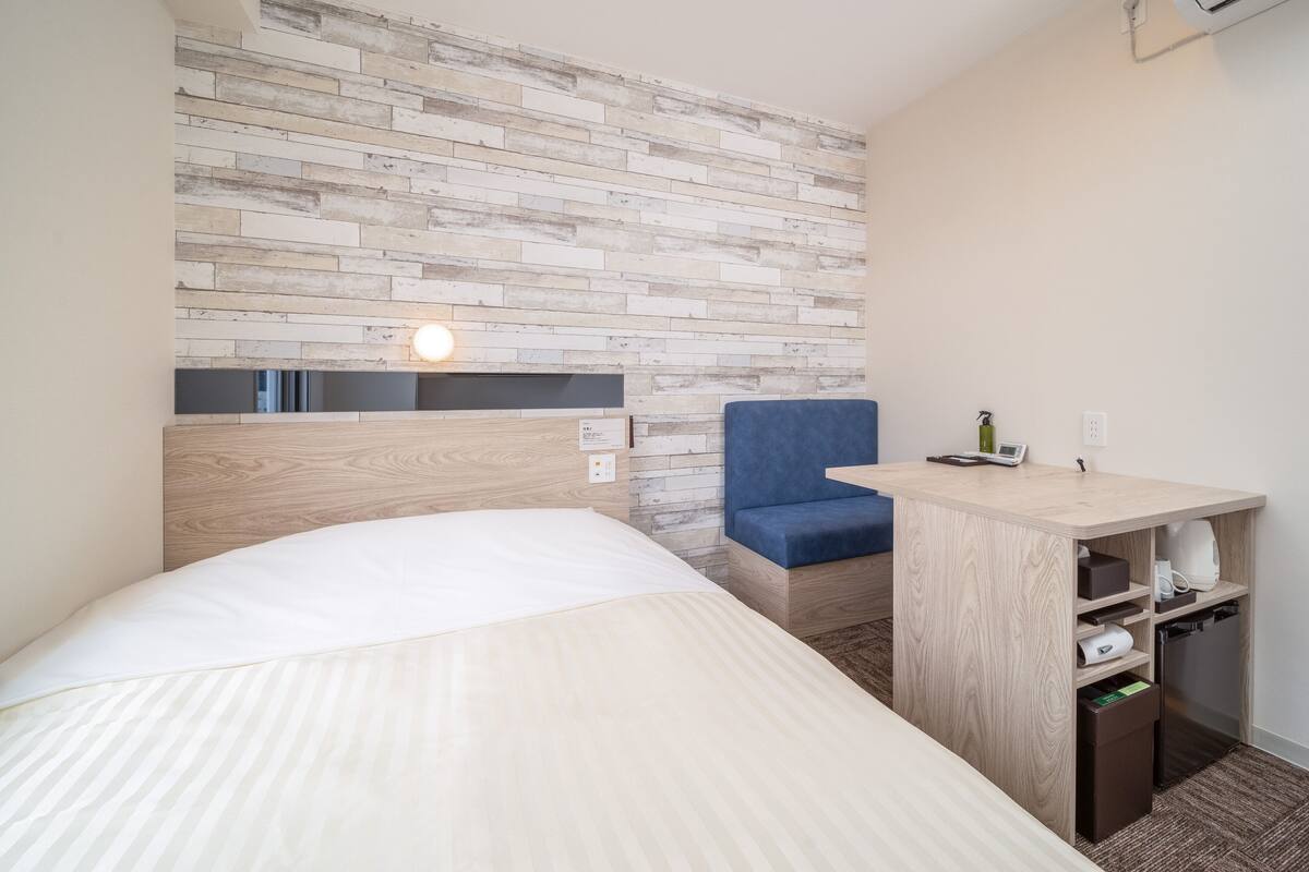 A hotel room with a bed, a desk with a chair, a wall-mounted air conditioner, and a built-in shelf with a trash bin.