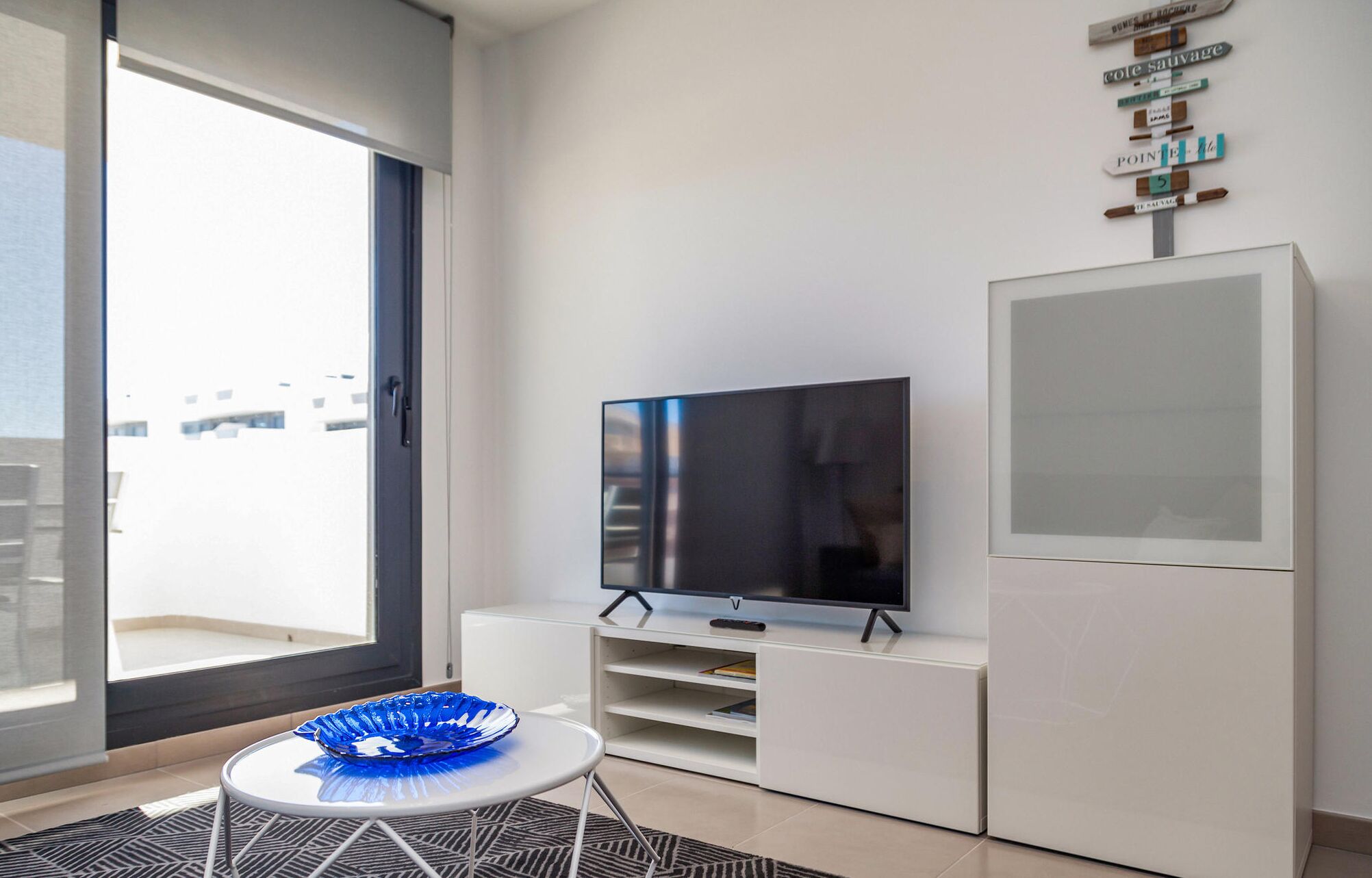 Ein modernes Wohnzimmer mit einem großen Flachbildfernseher auf einem weißen Standfuß, einer Glastür zu einem Balkon und einer blauen Dekorationsschale auf einem kleinen runden Tisch.