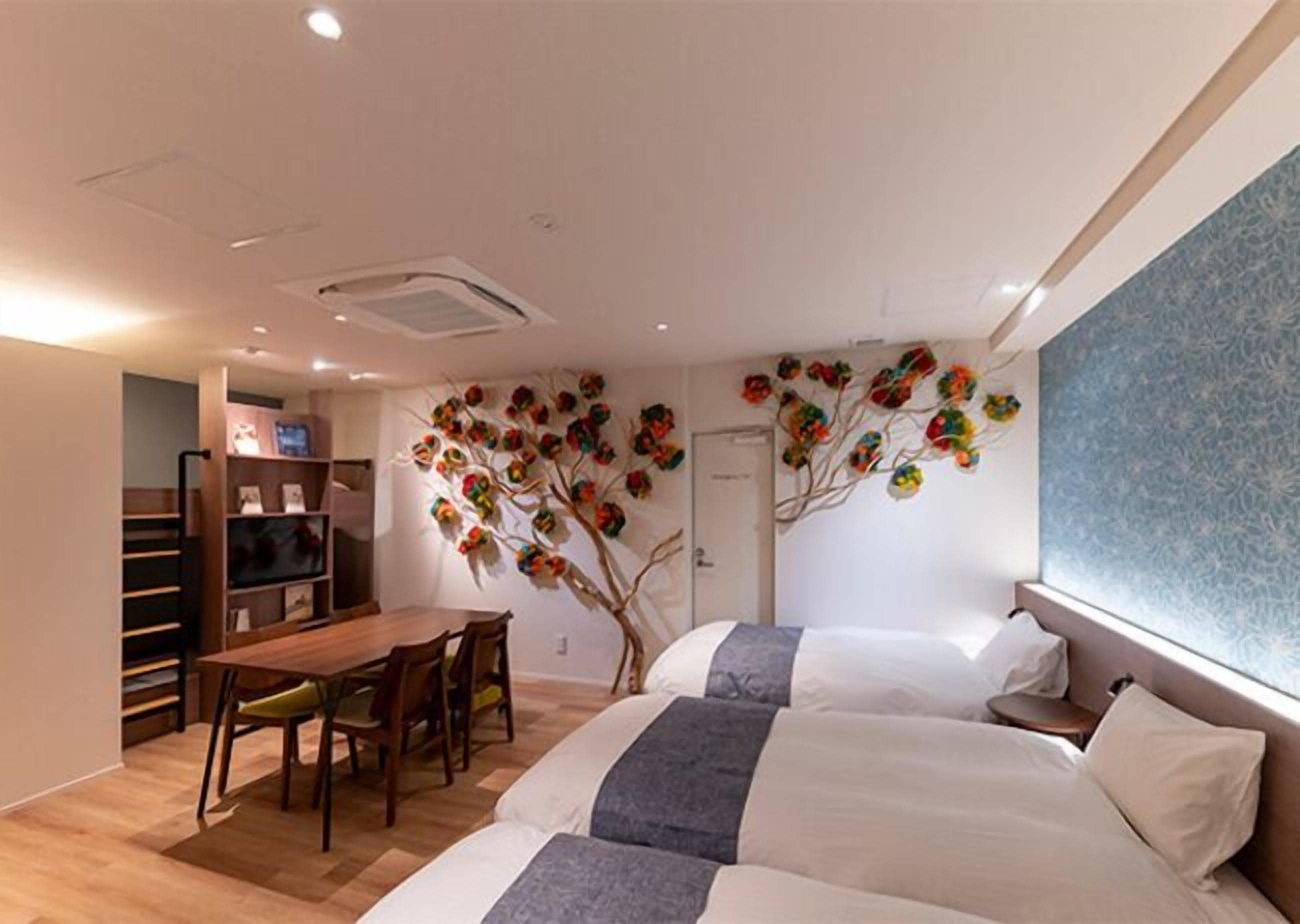 A hotel room with two beds, a dining table with chairs, a small kitchen area, and a decorative wall with a tree design.