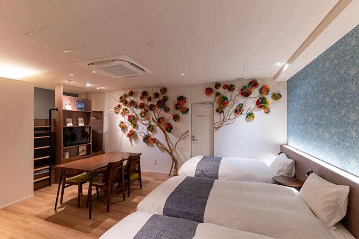 A hotel room with two beds, a dining table with chairs, a small kitchen area, and a decorative wall with a tree design.