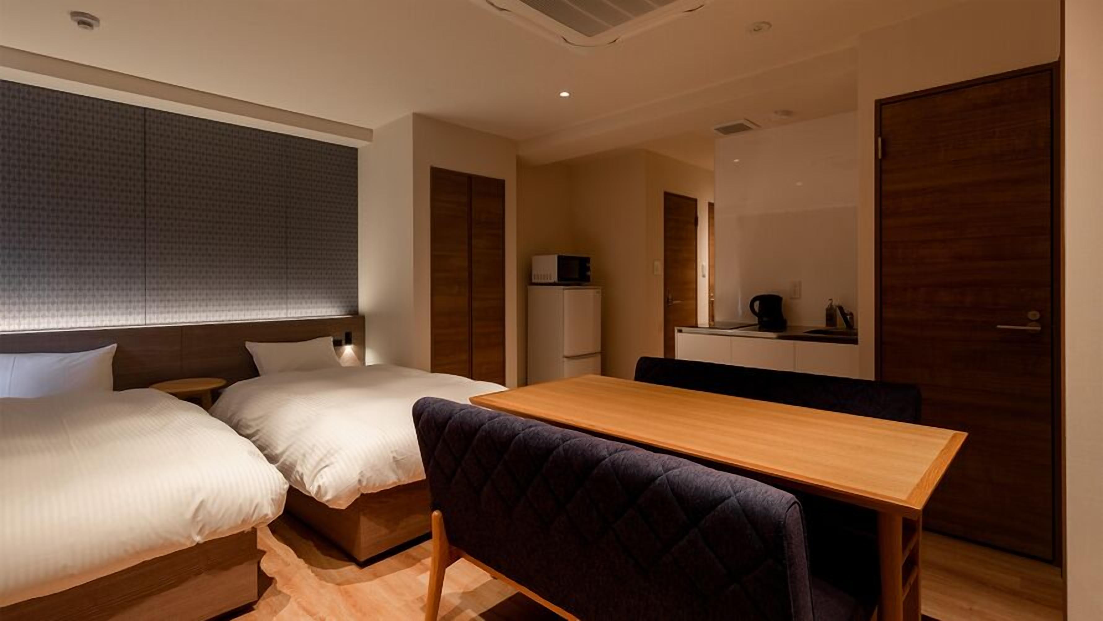 A hotel room with two beds, a wooden table, a small kitchenette, and a mirror.