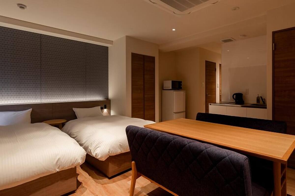 Ein Hotelzimmer mit zwei Betten, einem Holztisch, einer kleinen Küchenzeile und einem Spiegel.