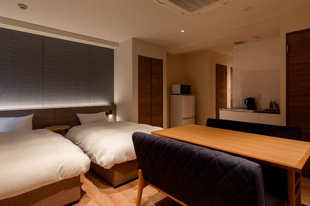 A hotel room with two beds, a wooden table, a small kitchenette, and a mirror.