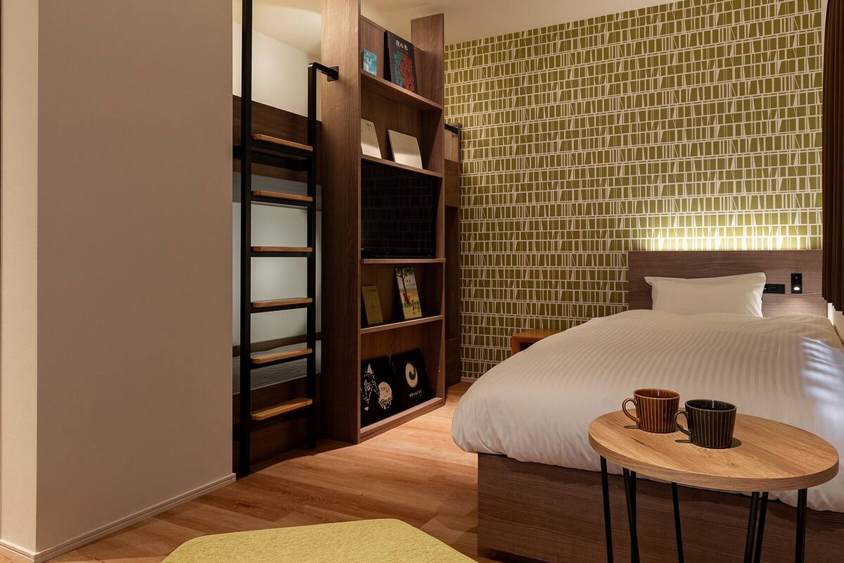 A hotel room with a bed, a wooden bookshelf, a small round table with two mugs, and a patterned wall.
