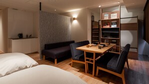 Quarto de hotel moderno com cama, sofá, mesa de jantar com cadeiras, estante e pequena cozinha equipada.