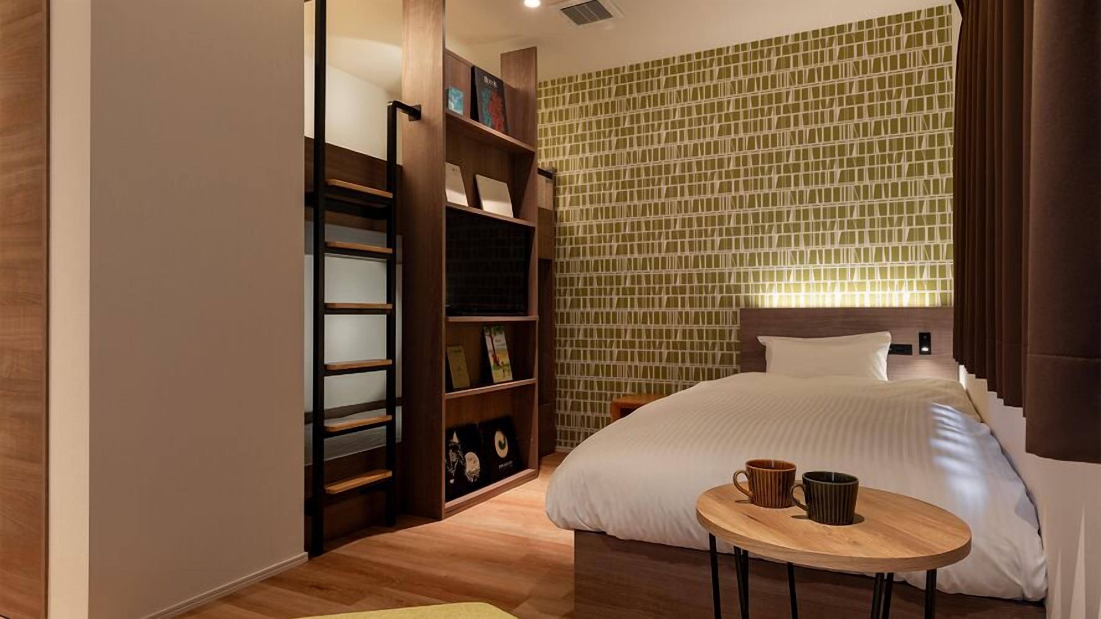 A hotel room with a bed, a wooden side table with two mugs, a bookshelf, and a patterned wall.