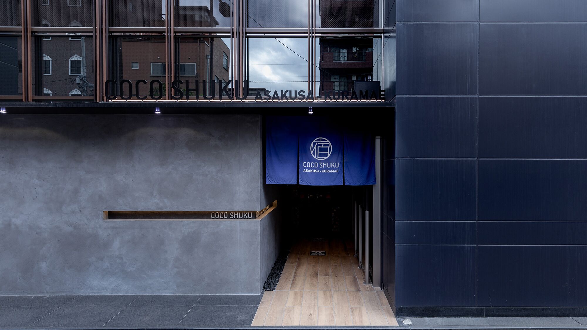 A modern building entrance with a sign reading "COCO SHUKU ASAKUSA-KURAMAe".