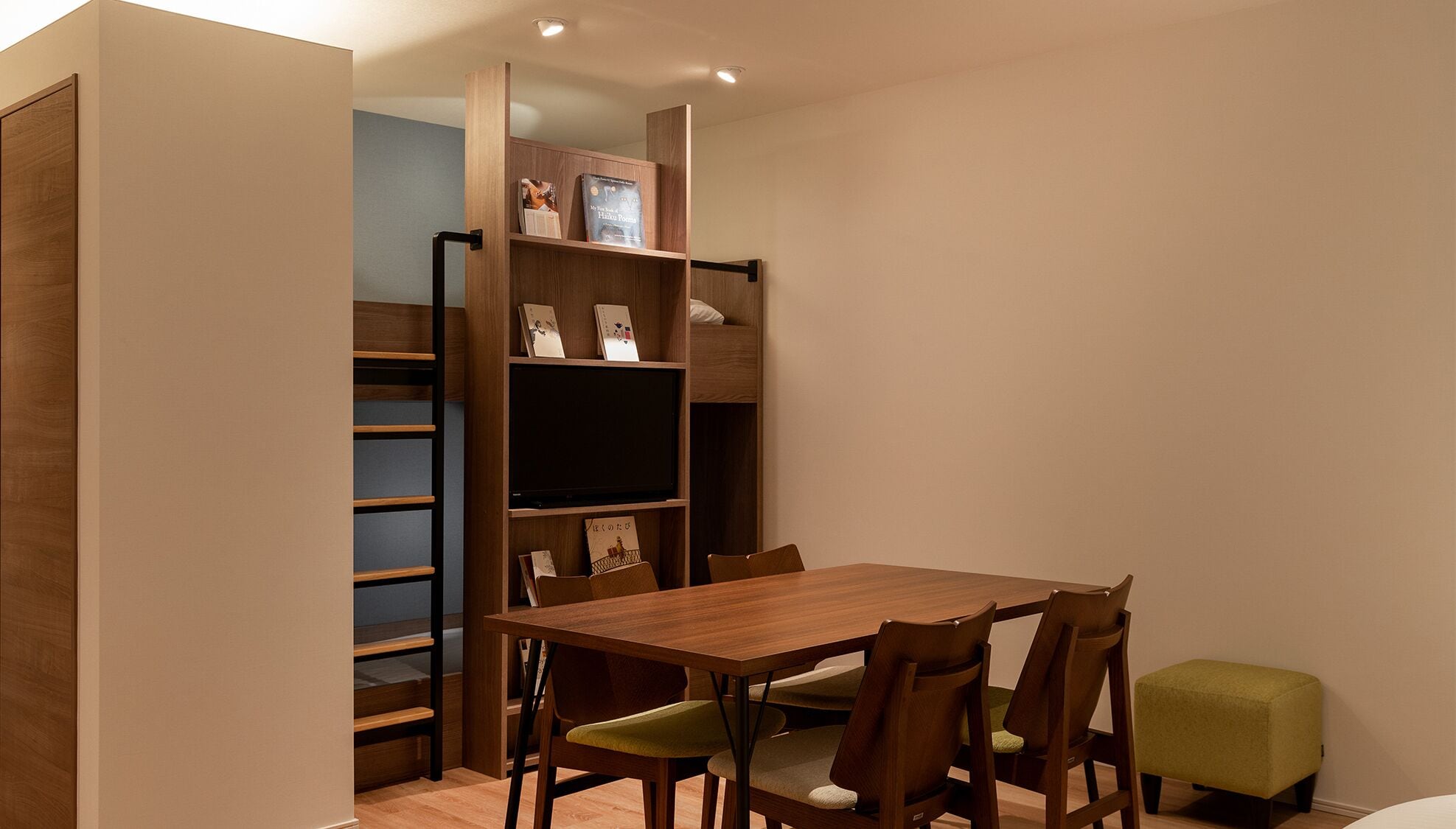 A compact room with a wooden table and chairs, a bunk bed, and a bookshelf with various items.
