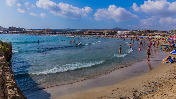 Une plage avec une eau d’un bleu limpide, des gens qui se baignent et prennent des bains de soleil, et des bâtiments en arrière-plan.