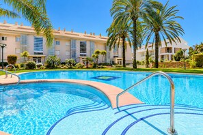 Une piscine à l’eau d’un bleu cristallin, entourée d’un jardin bien entretenu avec des palmiers, et un bâtiment doté de nombreux balcons en arrière-plan.