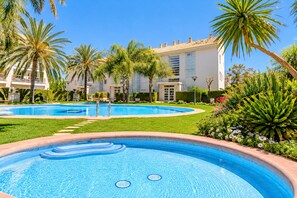 Una piscina con solárium alicatado, rodeada de palmeras y un edificio con techo de tejas.