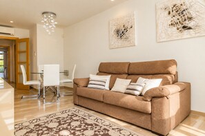 Een moderne woonkamer met een bruine bank, witte stoelen, een glazen salontafel en decoratieve wandkunst.