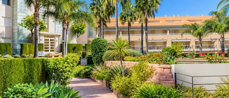 A landscaped pathway with palm trees and various shrubs, leading to a building with a tiled roof.