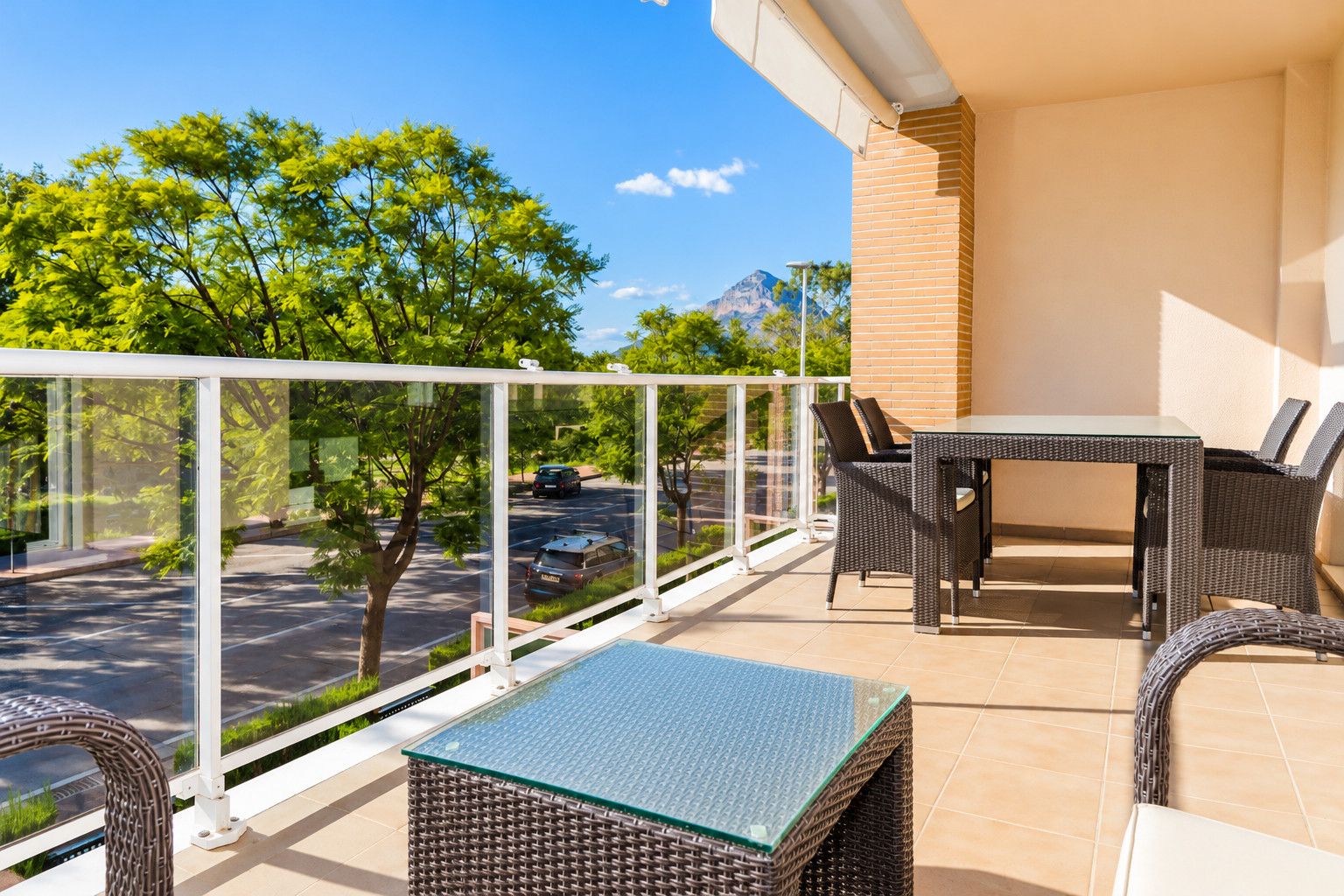 Un balcón con barandilla de vidrio, muebles de mimbre y vistas a una calle y montañas.