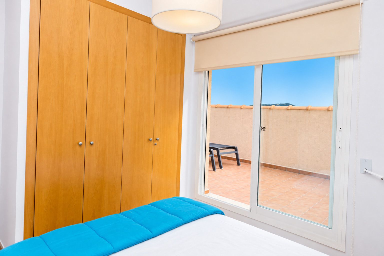 Ein Schlafzimmer mit einem Holfischschrank, einem Bett mit blauer Matratze, einem Fenster mit Blick auf eine Terrasses und einer Deckenleuchte.