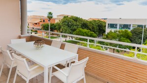 Un balcon avec une table et des chaises blanches, un vase décoratif et une vue sur des immeubles et des arbres.