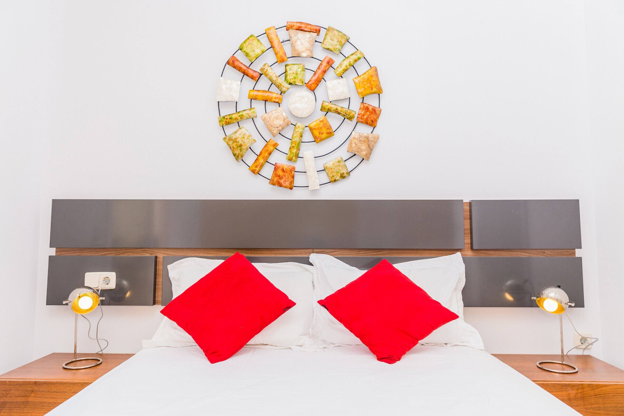 Un lit bien fait, agrémenté d’oreillers rouges, une œuvre d’art circulaire accrochée au mur et des lampes de chevet.