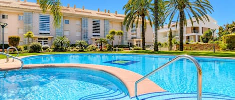 En swimmingpool med krystalklart blåt vand, omgivet af en velplejet have med palmer og en bygning med flere balkoner i baggrunden.