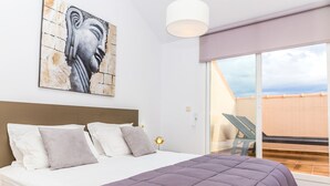 Une chambre à coucher avec un lit, des oreillers, une tête de lit, une œuvre d’art accrochée au mur, une lampe de chevet et une vue sur l’extérieur.