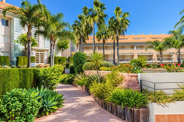 A landscaped pathway with palm trees and various shrubs, leading to a building with a tiled roof.
