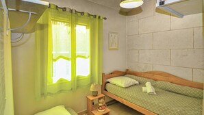 A small, cozy room with two single beds, a wooden headboard, a small bedside table with a lamp, and a window with sheer curtains.