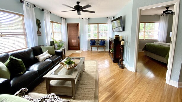 A living room with a black leather sofa, a coffee table with plants, a wooden floor, a ceiling fan, and a TV mounted on the wall. - The Tropical Bungalow (Safety Harbor)
