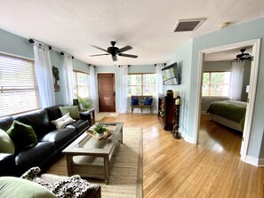 A living room with a black leather sofa, a coffee table with plants, a wooden floor, a ceiling fan, and a TV mounted on the wall. - The Tropical Bungalow (Safety Harbor)