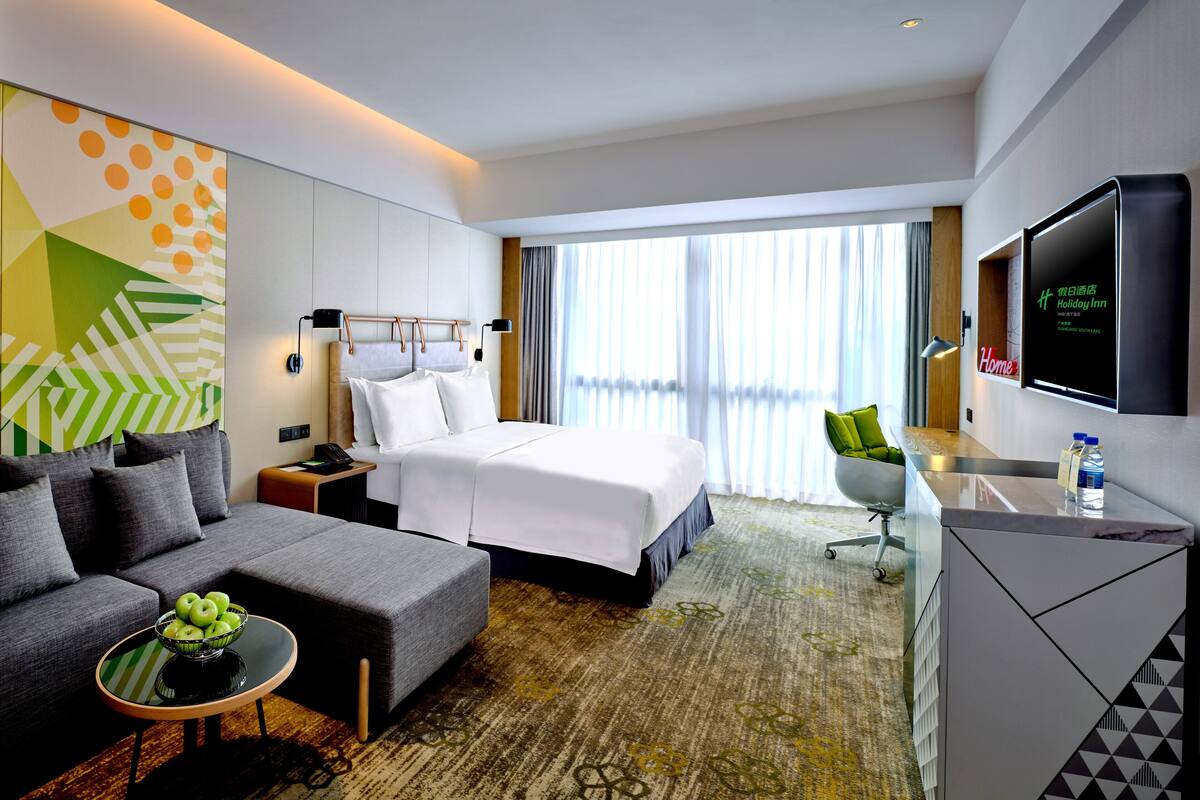 A hotel room with a bed, a sofa, a desk, a TV, and a large window with curtains.