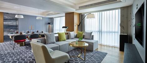 A modern hotel room with a dining area, a sofa set, a TV, and a large window with curtains.