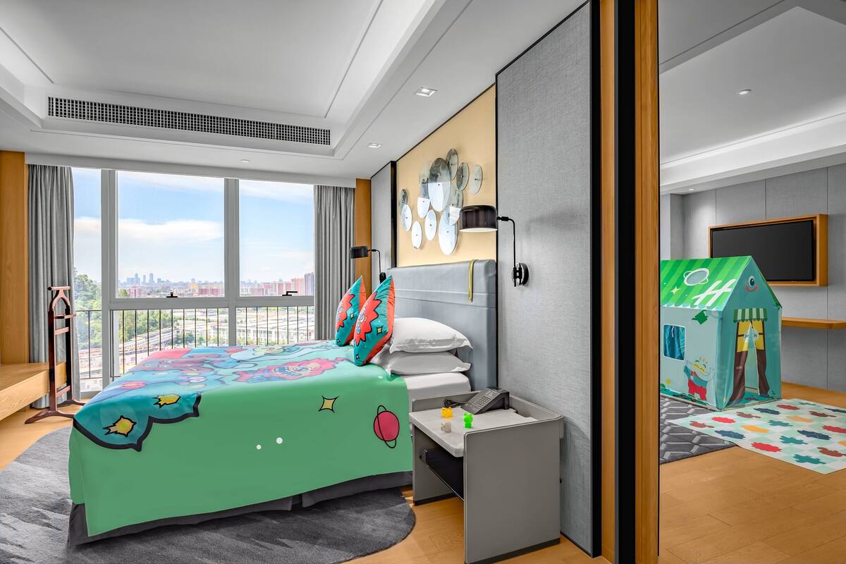 A modern hotel room with a large bed, a desk, a playhouse, and a city view.