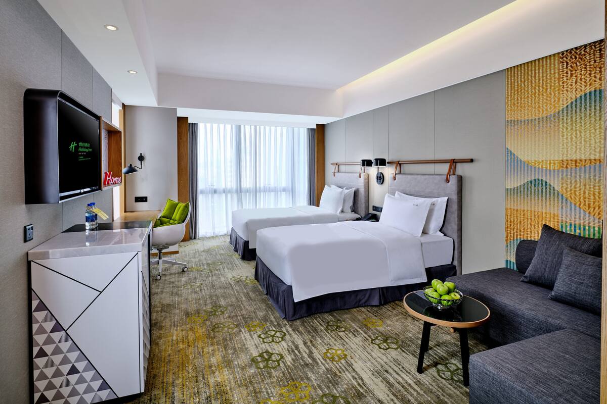 A hotel room with two beds, a desk, a sofa, a TV, and a window with curtains.