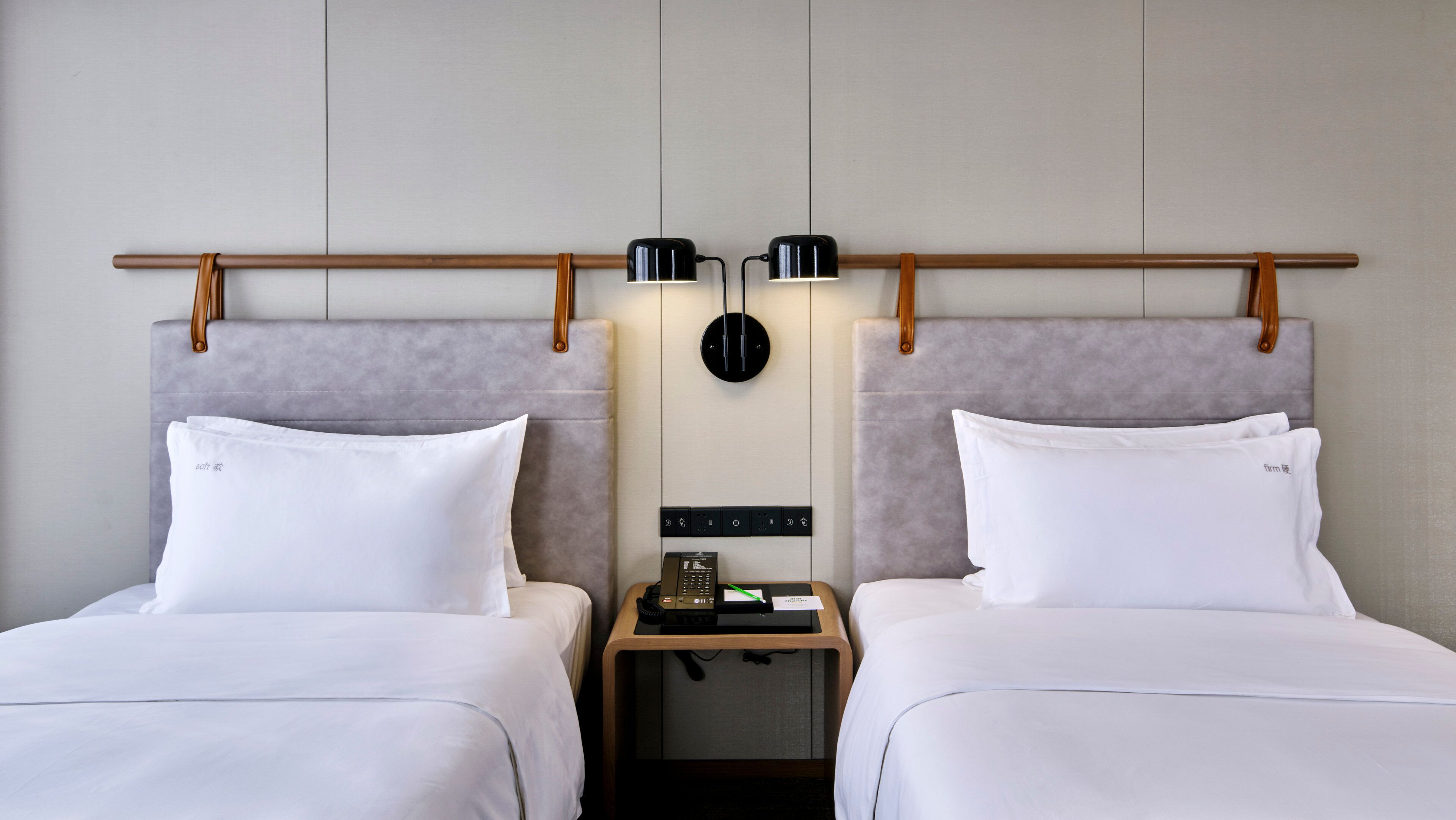 Een hotelkamer met twee bedden, een houten hoofdbord, een nachtkastje met telefoon en notitieboekje, en een wandlamp.