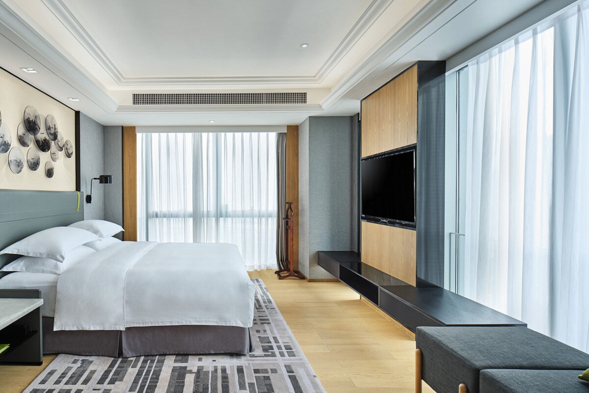 A modern hotel room with a large bed, a TV, and a large window with sheer curtains.