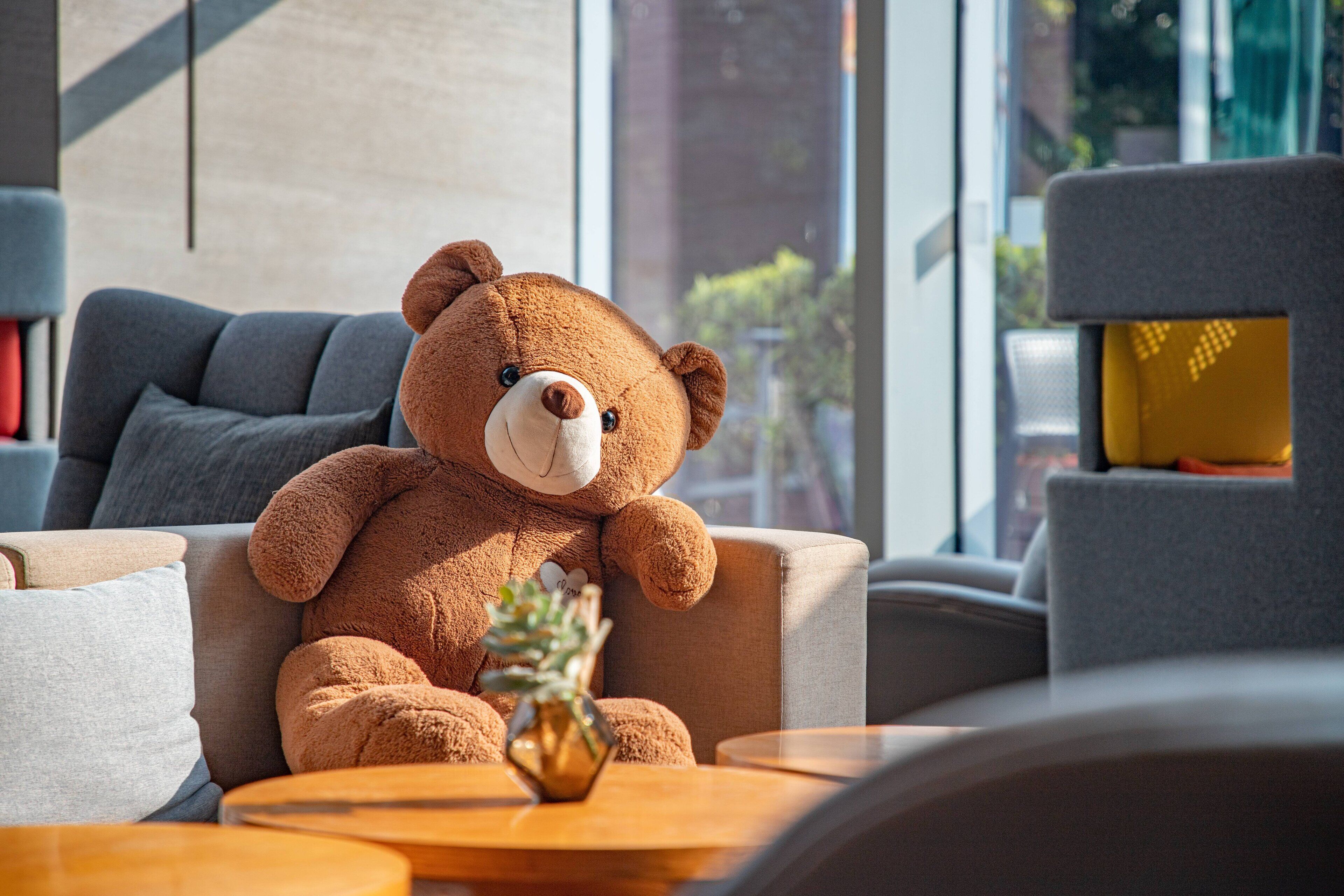A plush brown teddy bear sitting on a grey sofa in a modern living room with a small table and a vase of flowers.