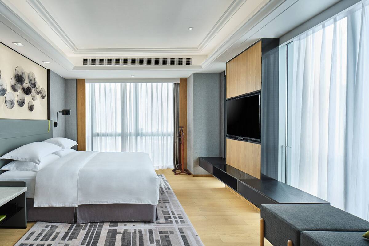 A modern hotel room with a large bed, a TV, and a large window with sheer curtains.