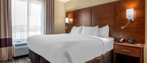 A hotel room with a large bed, a bedside table with a phone and a coffee maker, a wall-mounted lamp, and a window with curtains.