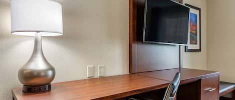 A hotel room with a wooden desk, a chair, a lamp, a flat-screen TV mounted on the wall, and a framed picture.