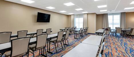 Una sala de conferencias con varias mesas y sillas, un televisor de pantalla plana montado en la pared y ventanas amplias con cortinas.