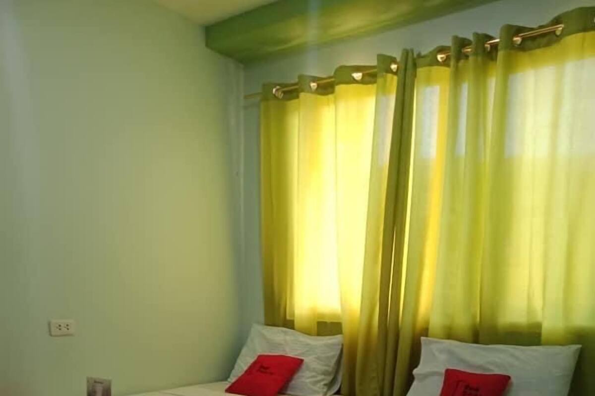Hotelkamer met twee bedden, elk voorzien van een rode kussen en een witte dekbedovertrek. Er is een raam met gele gordijnen, en de wand is groen.