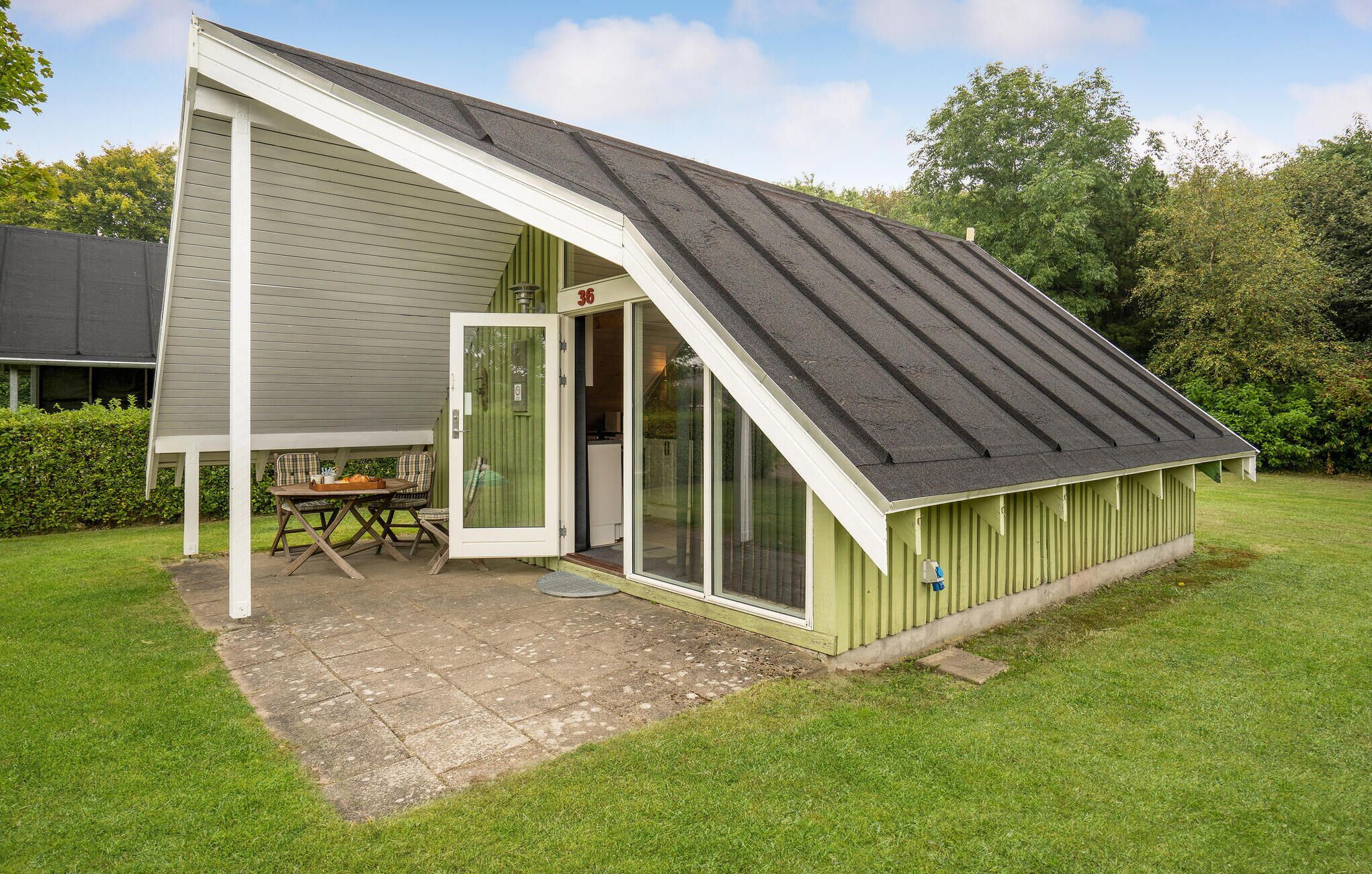A small, single-story building with a sloped roof, a covered patio with a table and chairs, and a door leading to an interior space.