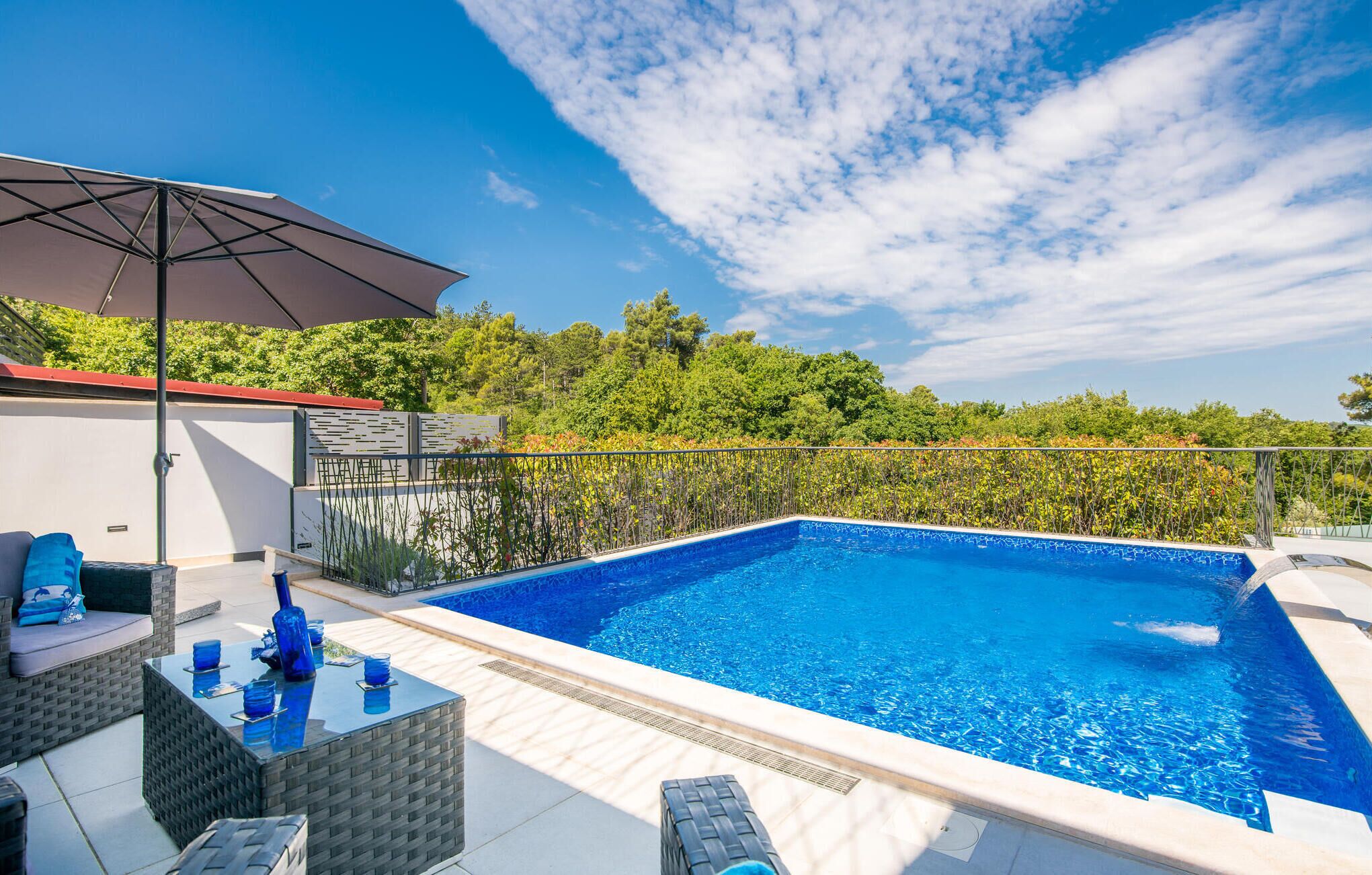 A swimming pool with a tiled deck, outdoor furniture, and a pergola providing shade.