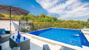 Ein Swimmingpool mit einer gefliesten Terrasse, Gartenmöbeln und einer Pergola, die Schatten spendet.