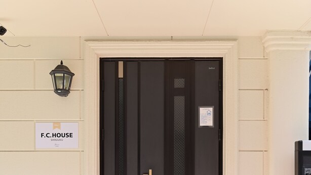 Eine schwarze Tür mit goldener Klinke, ein Schild mit der Aufschrift "F.C.HOUSE SHINJUKU" und ein schwarzer Laternenaufsatz an der Wand.