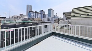 Eine Dachterrasse mit weißer Brüstung, umgeben von Gebäuden, darunter ein hoher, blauer Wolkenkratzer und ein weißes Gebäude mit geneigtem Dach.